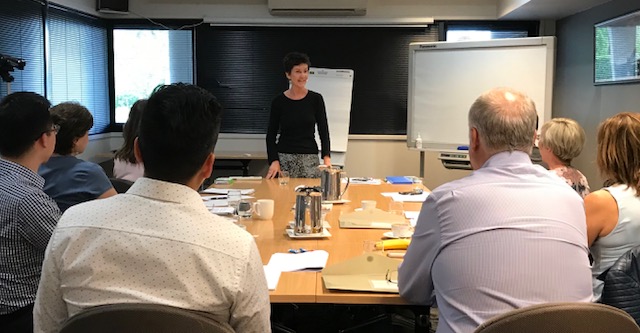 Sarah Denholm standing in front of a table training a small group class in the complete presentation skills course melbourne