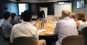 Sarah Denholm standing in front of a table training a small group class in the complete presentation skills course melbourne